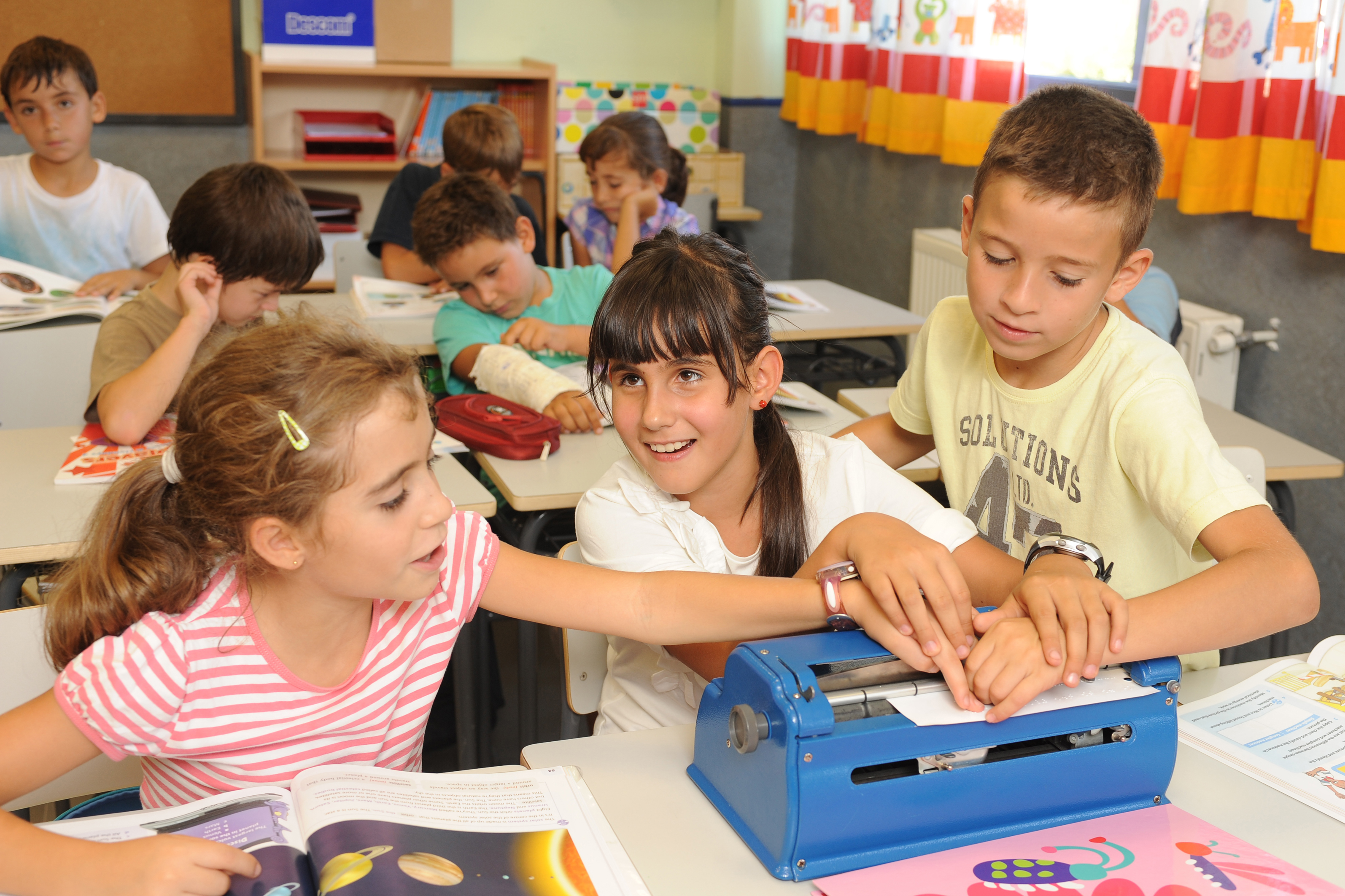 Alumna amb una màquina d'escriptura en braille, encoltada per dos companys a l'aula.